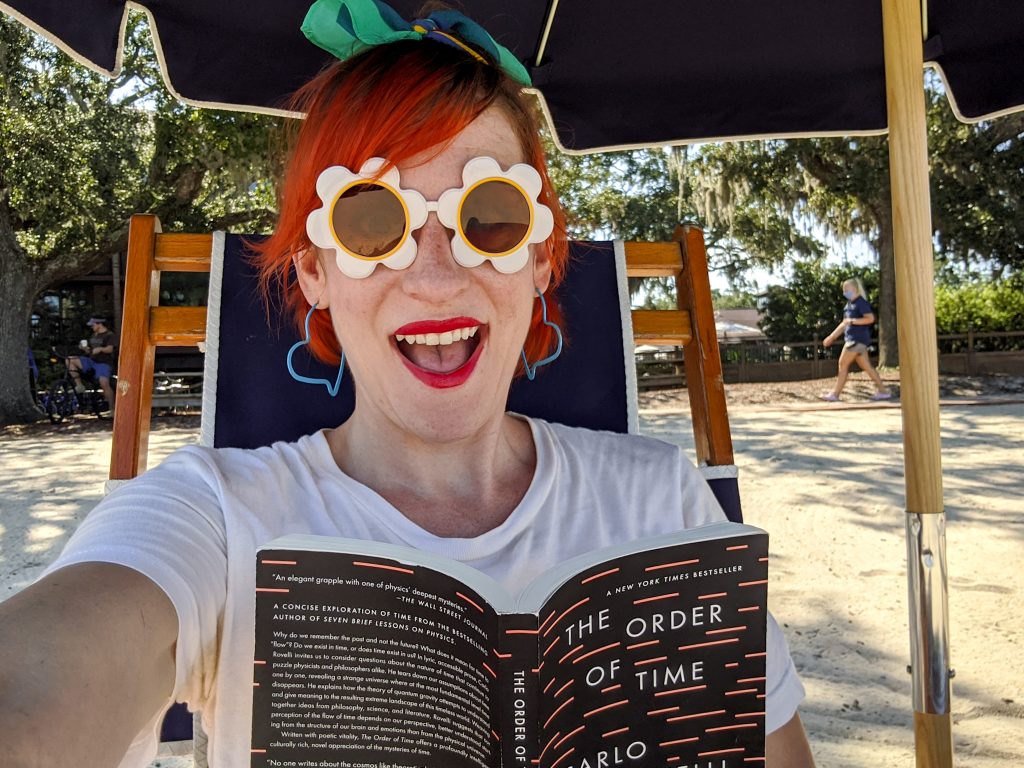 A redheaded woman in flowered sunglasses reads a book on the beach