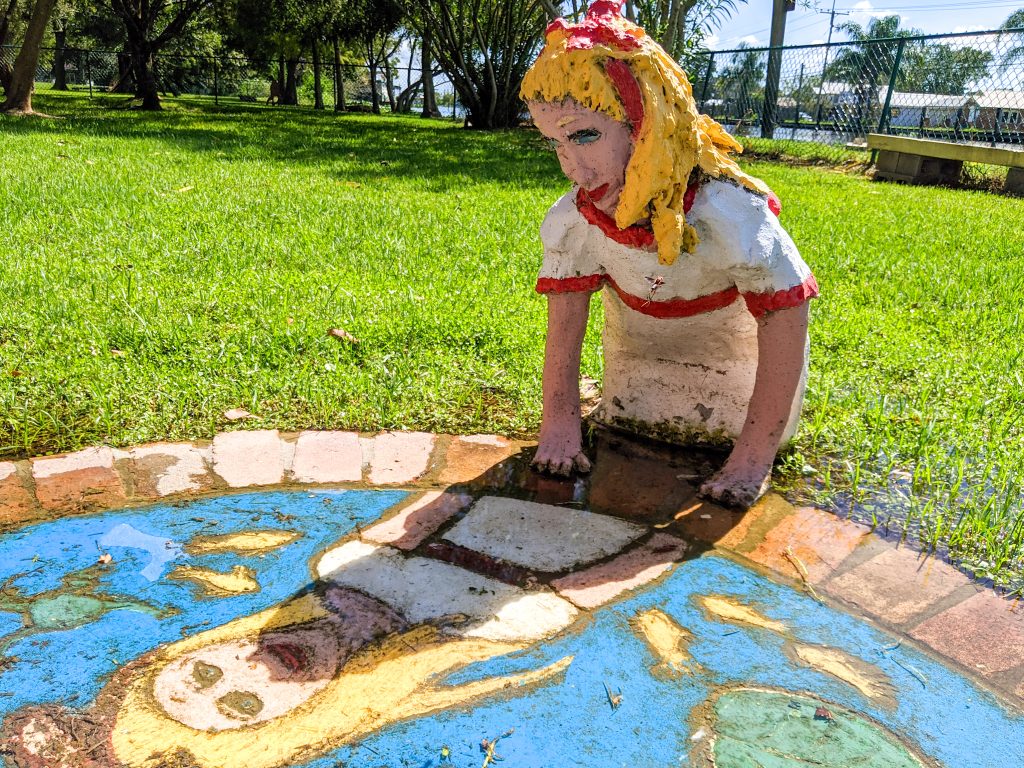A folk art sculpture of a blonde girl regards her own reflection in a painting of a pond. The sculpture garden is one of several easy day trips out of New Orleans. 
