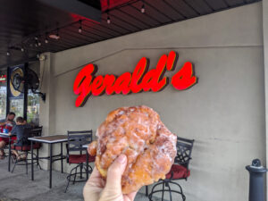 A hand holds up a large apple fritter in front of a red sign reading "Gerald's"