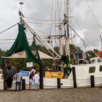 People line up at a white shrimp boat docked under a stormy sky