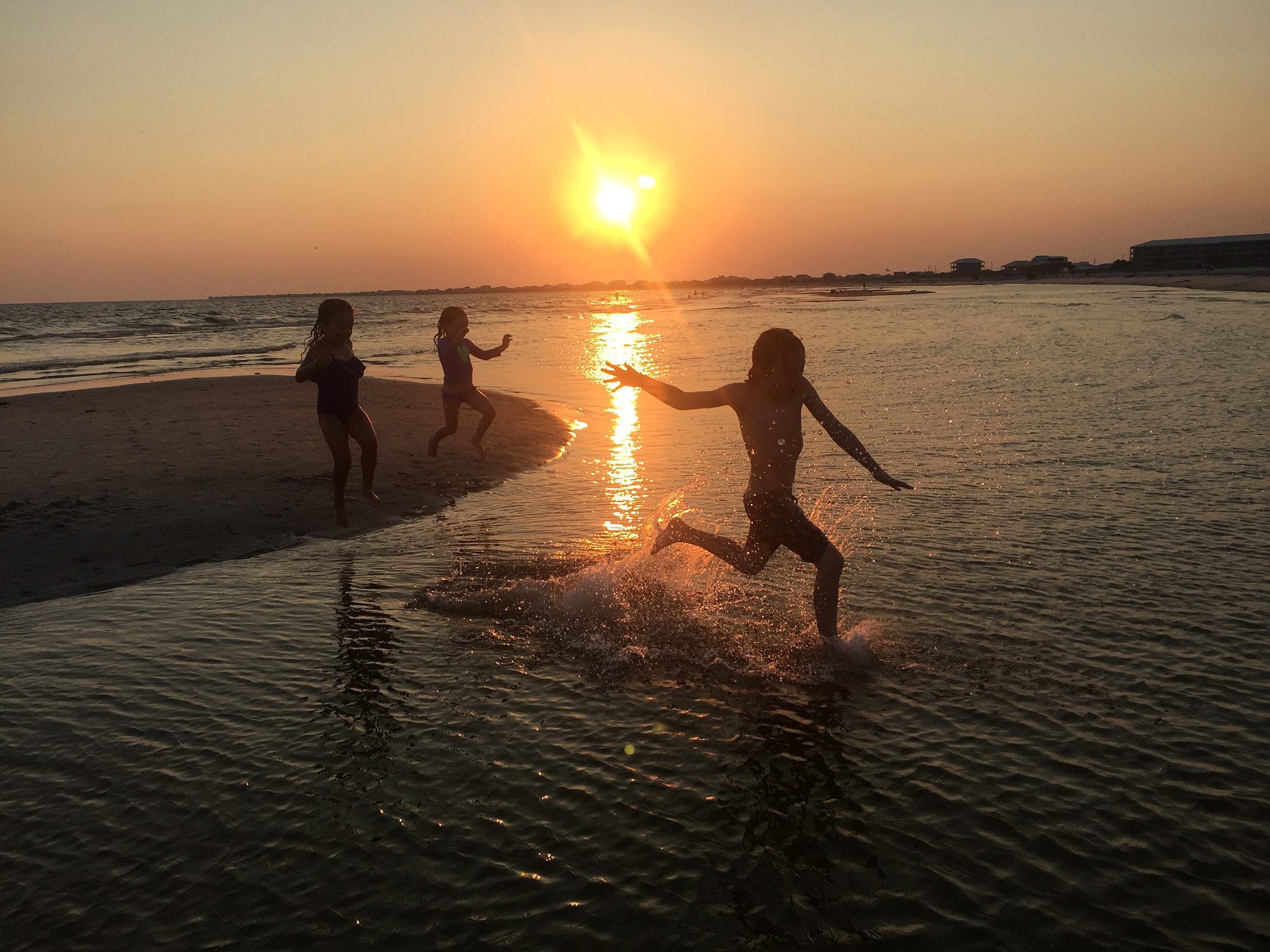 Three children run along a beach at sunset.